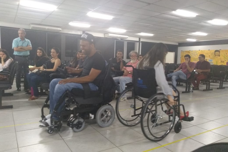 #pratodosverem imagem que registra o momento de uma dinâmica realizada no auditório. Ao centro, um homem e uma mulher, de costas um para o outro, ambos em cadeiras de rodas. À direita, pessoas na plateia observam a atividade. Fim da descrição.