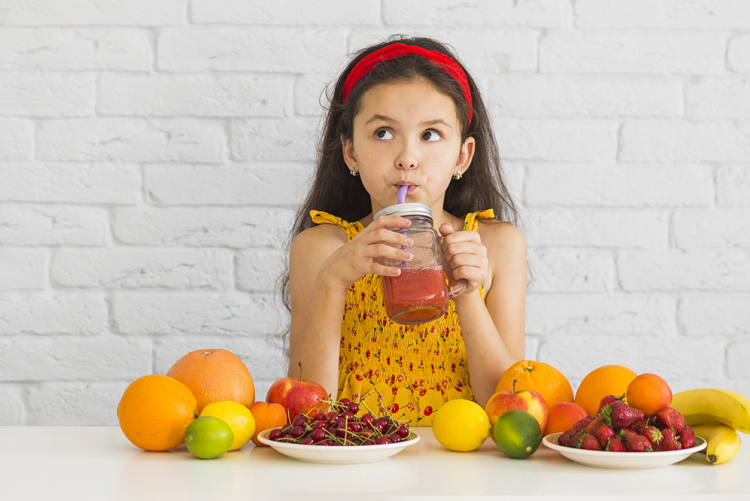 Menina de vestido amarelo tomando suco no canudo com mesa cheia de frutas a sua frente