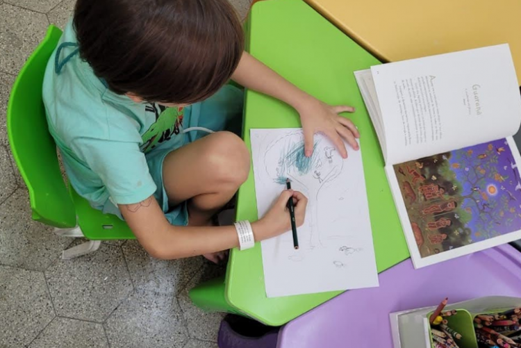 Foto de menino sentado numa cadeira verde e desenhando sobre uma mesa verde també
