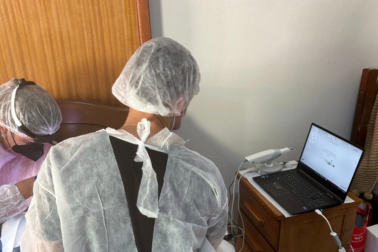 Dois profissionais da odontologia com touca e capa protetora, de costas, manipulando um paciente enquanto observam à direita a tela de um laptop