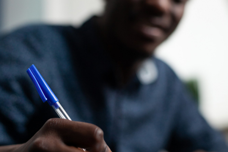 Rapaz negro de camisa azul, escrevendo em um caderno com uma caneta azul