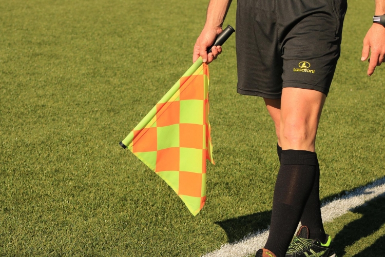 Pernas de um juiz em um campo de futebol, em deslocamento. Também se vê a mão direita segurando uma bandeira.