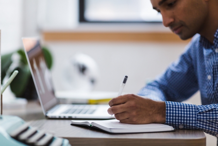 Homem com laptop aberto anotando algo em um caderno