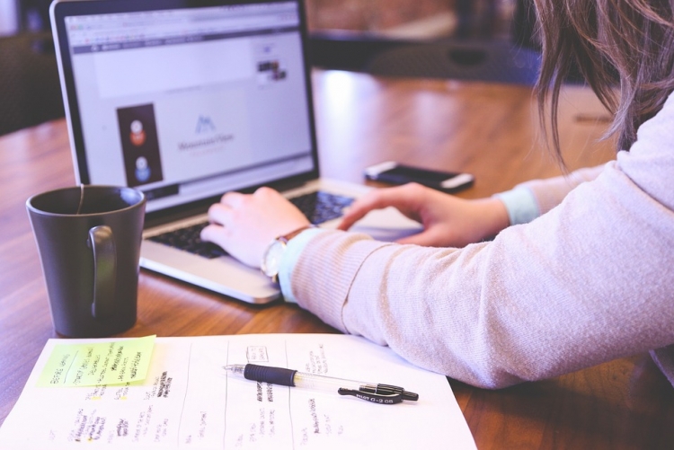 Imagem: mulher digitando no notebook com caneca e caderno de anotações ao lado