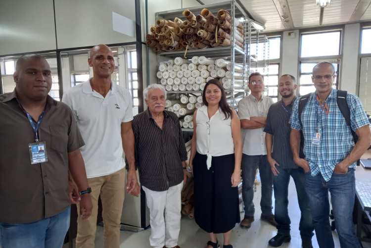 Foto na sala da SAEP onde ao fundo se encontra uma estante com rolos de plantas baixas de obras. Na frente da estante, estão os seguintes servidores da esquerda para a direita: Mario Vinicius Campos, arquivista; Claudio Muniz, arquivista e conselheiro do CONARQ, Julio Rogerio, Superintendente da SAEP; Camilla de Aquino, arquivista e Coordenadora da CAR; Jose Luiz Vieira, servidor da SAEP; Felipe Chagas, arquivista da CAR e Marcos Vinicius Andrade,  Assistente da SDC