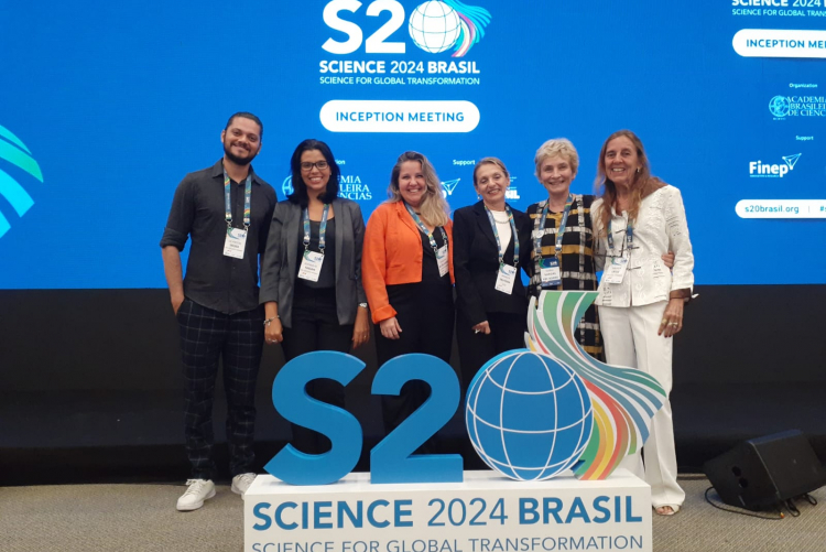 Foto dos representantes da UFF no evento do S20. Os pesquisadores da universidade, Da esquerda para a direita: Ueverton Souza, Joanna Fabja, Thaiane de Oliveira, Monica Savedra, Maria Vargas e Andrea Latge, estão de pé na frente da placa com o logotipo do evento