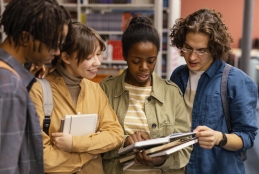 Foto de 4 estudantes universitários negros e brancos, em pé, fazendo a leitura de um livro