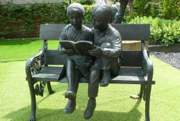 Escultura de duas crianças lendo um livro em uma cadeira, dentro de um jardim
