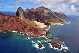 #ParaTodosVerem A imagem mostra um conjunto de montanhas na Ilha da Trindade, parte do arquipélago Trindade e Martim Vaz. A ilha é cercada por um mar azul-esverdeado e no alto da imagem observa-se um céu azul-celeste repleto de nuvens
