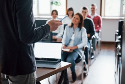professor em pé em sala de aula com computador e alunos sentados em cadeiras