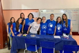 Professora rodeada de alunos vestidos com uniforme azul que identifica o projeto em frente a uma mesa