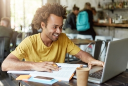 Estudante universitário negro sorrindo e sentado na frente de um computador com livros na mesaros