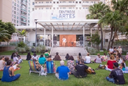 várias pessoas sentadas de costas nos jardins da reitoria da UFF, assistindo a uma apresentação musical