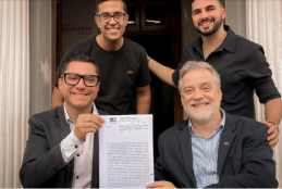 foto de dois homens sentados segurando uma escritura sorrindo. Ao fundo, outros dos homens em pé sorrindo compõe a foto.