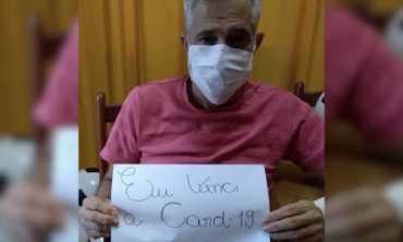 homem de camisa rosa, de máscara branca, sentado em cadeira de rodas, segurando um papel com os dizeres: eu venci a Covid-19
