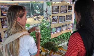 Mulher branca, cabelo loiro, segurando um pote com planta, fazendo explanação para outra mulher branca, de cabelos pretos