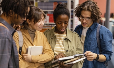 Foto de 4 estudantes universitários negros e brancos, em pé, fazendo a leitura de um livro