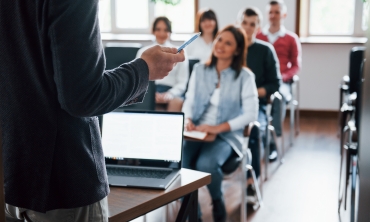professor em pé em sala de aula com computador e alunos sentados em cadeiras