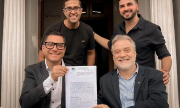 foto de dois homens sentados segurando uma escritura sorrindo. Ao fundo, outros dos homens em pé sorrindo compõe a foto.